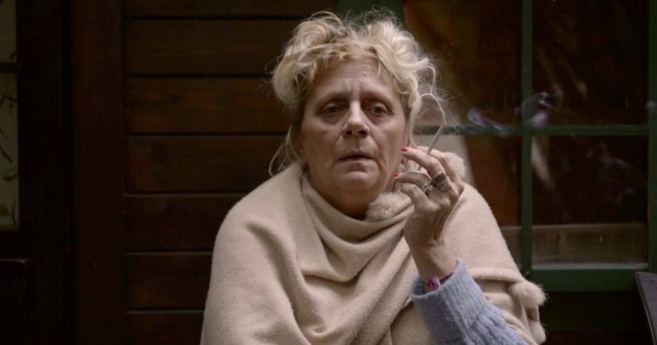 Une femme se tient devant une maison, cigarette à la main, profitant d'un moment de calme à l'extérieur.