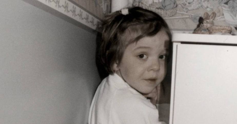 Une jeune enfant jette un coup d'œil malicieux depuis l'arrière d'un cabinet, affichant une expression triste.